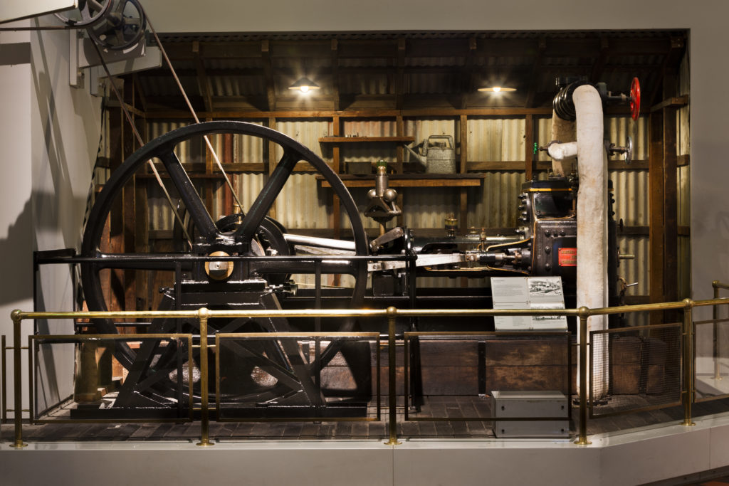 A Museum exhibit of a steam engine. It consists of a large flywheel attached to two pulleys. The engine sits on a large platform, bordered by a railing. Behind it is a mock engine shed wall with corrugated iron and an oil can sitting on a shelf. 